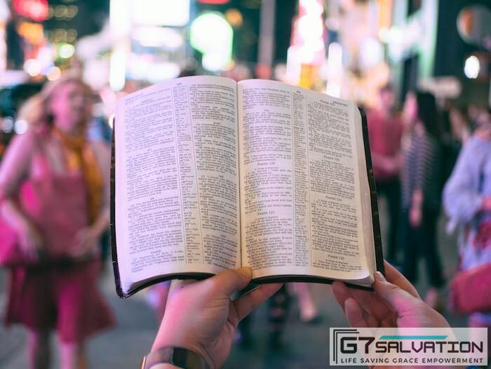 person holding bible on road with people walking on sidewalk beside buildings during nighttime